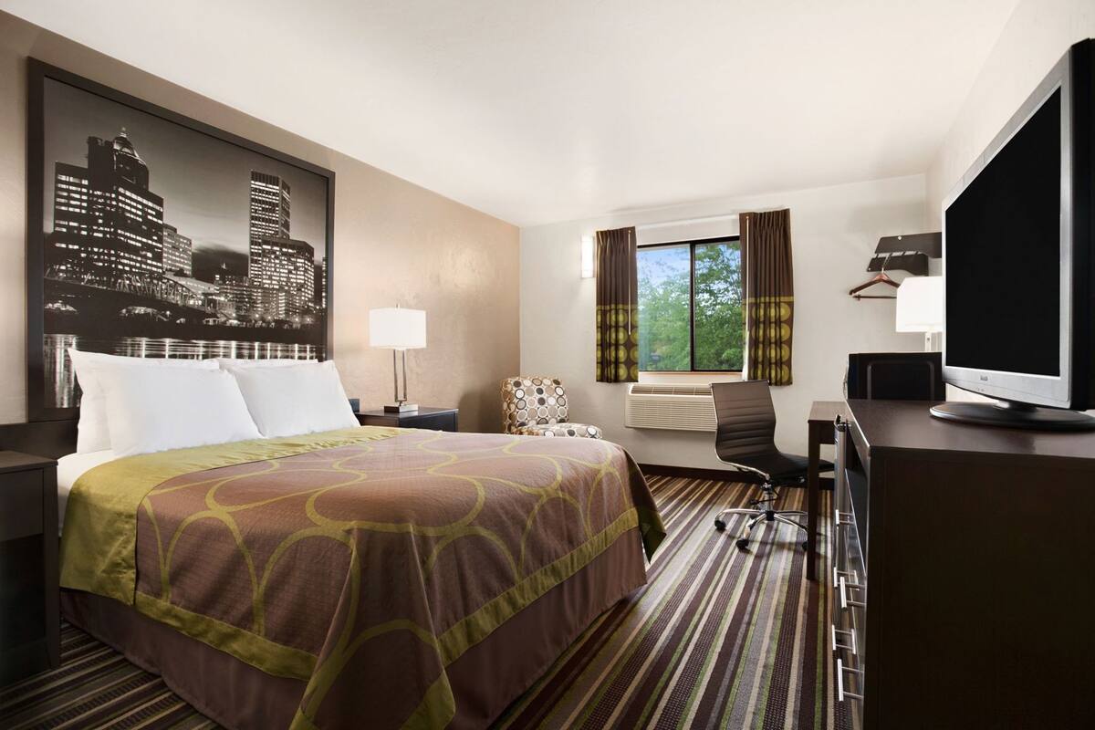 Een hotelkamer met een bed, een bureau met een televisie, een stoel, een raam met gordijnen en een stadsgezichtschilderij aan de muur.