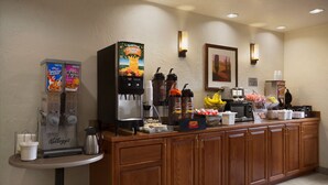 Un mostrador de desayuno de hotel con una máquina de café, un dispensador de zumo y una variedad de alimentos.