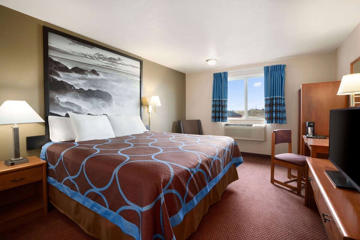 A hotel room with a bed, a desk, a chair, a TV, a window with curtains, and a painting on the wall.