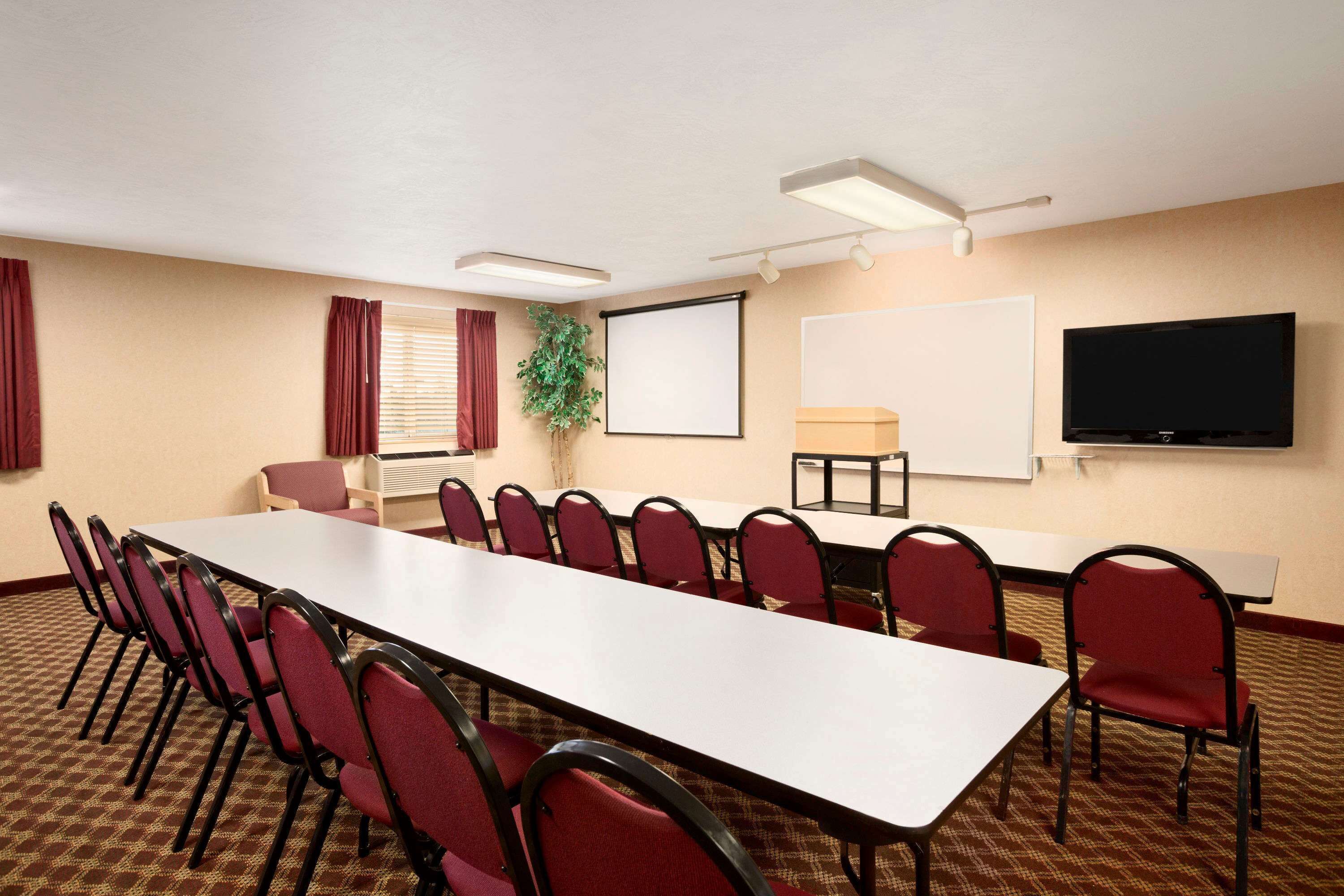 Una sala de conferencias con una larga mesa, sillas rojas, una pantalla de proyector, un televisor y una planta.