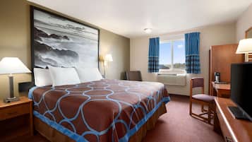 Habitación de hotel con una cama grande, un escritorio con televisor, una silla, una lámpara de escritorio y un ventanal con cortinas.