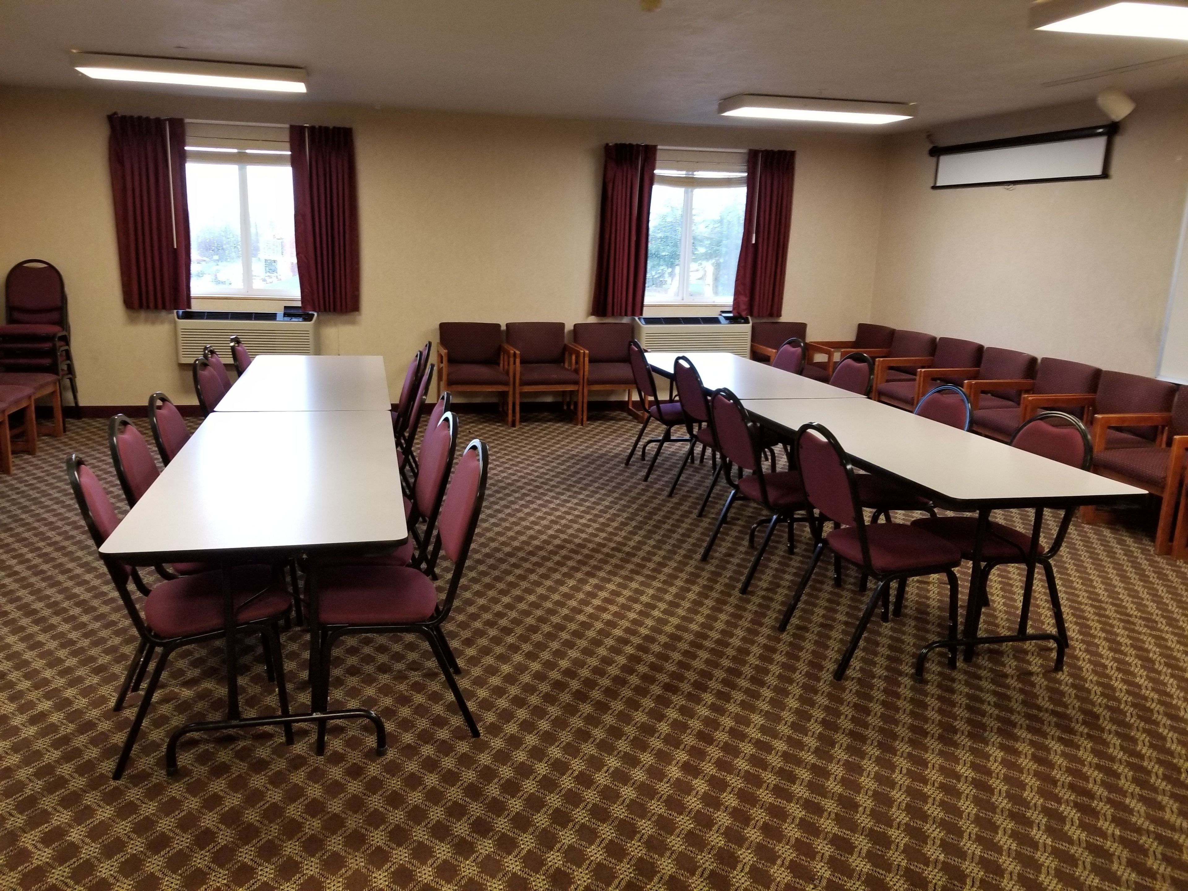 A conference room with a long table and chairs, air conditioning units, windows with curtains, and a projector screen on the wall.