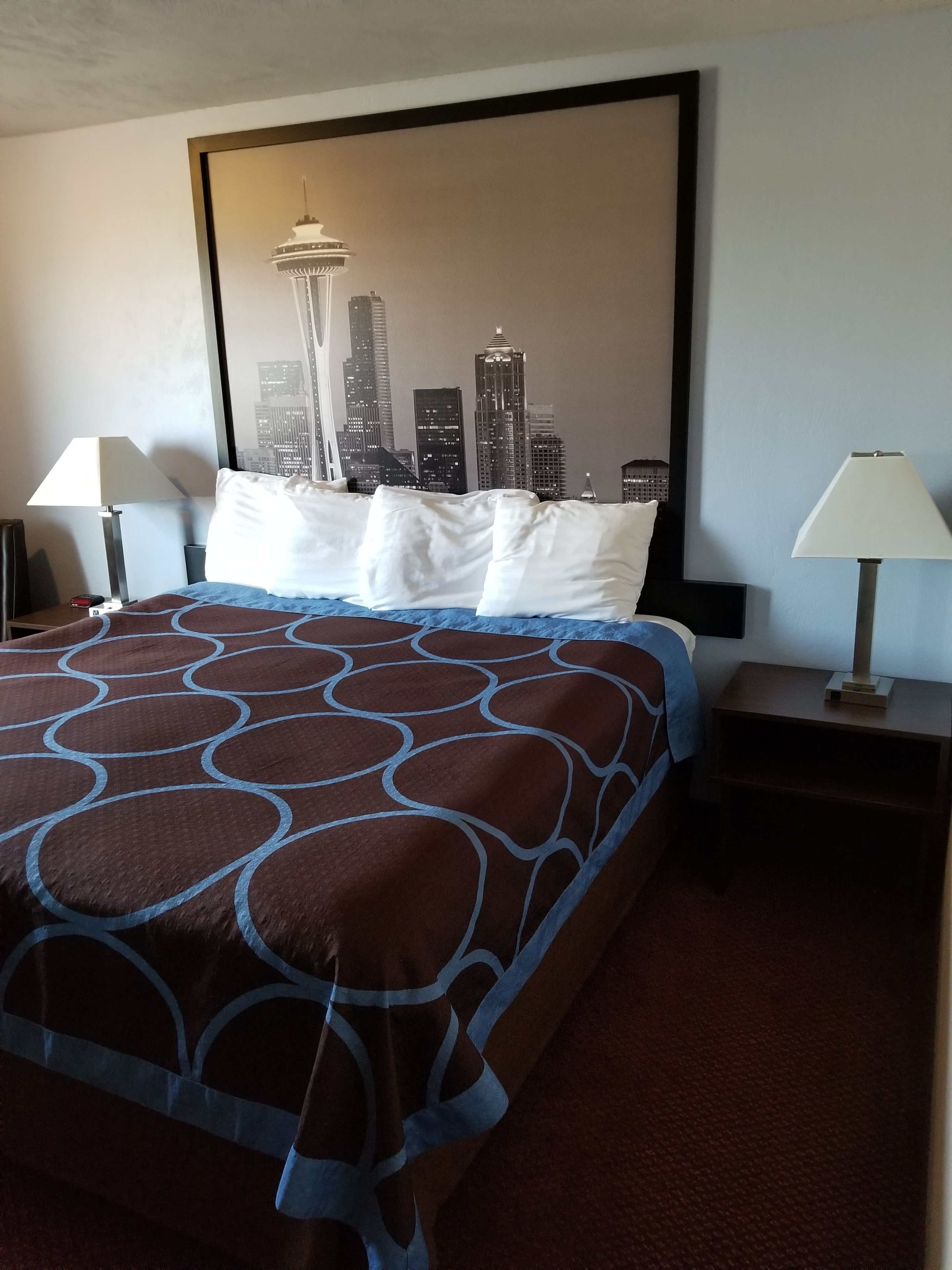 Habitación de hotel con una cama, dos mesitas de noche, una lámpara a cada lado y un gran cartel con un paisaje urbano en la pared.