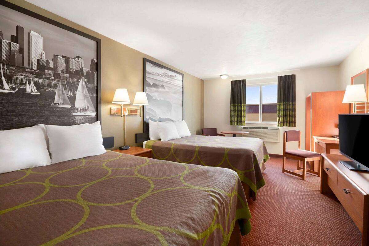 A hotel room with two beds, a desk with a TV, a chair, a window with curtains, and artwork on the walls.