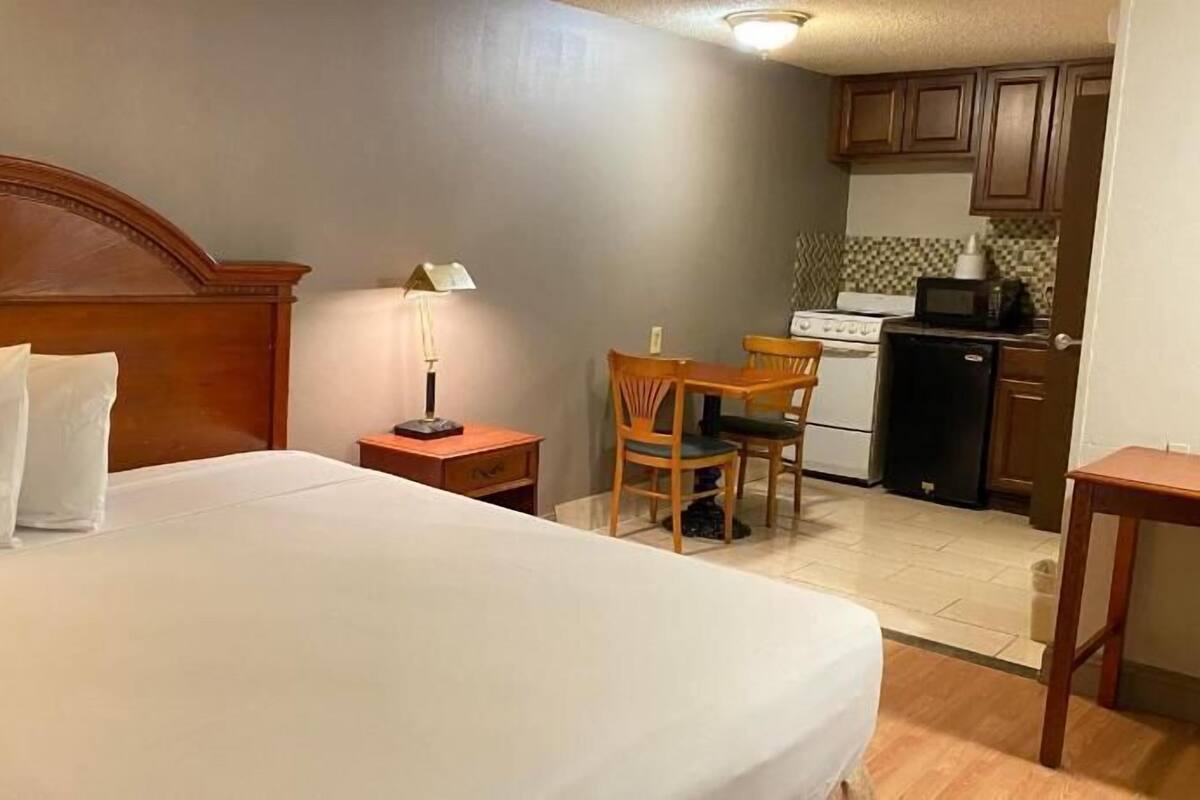 A hotel room with a bed, a nightstand with a lamp, a wooden chair, a small table, and a kitchenette with cabinets and a stove.