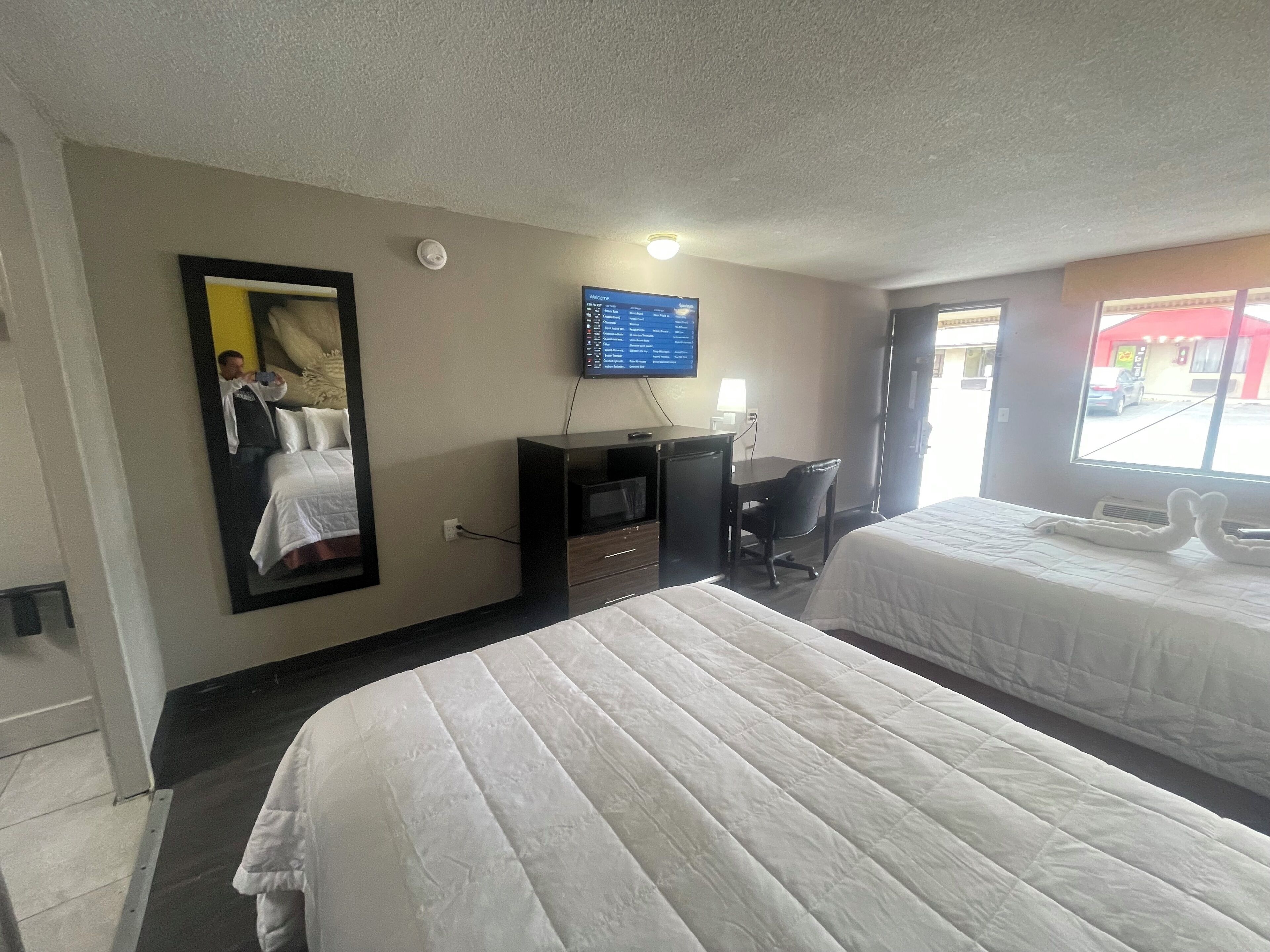 A hotel room with two beds, a desk, a TV, and a window with a view of the outside.