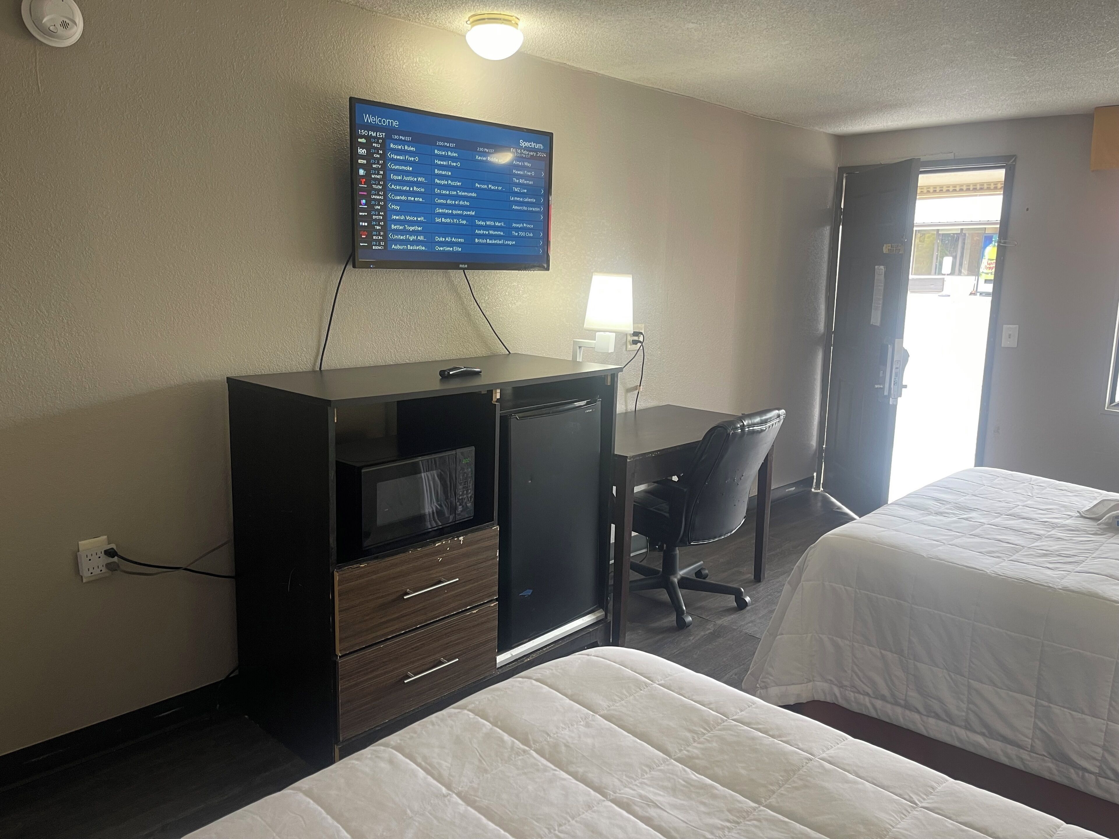 A hotel room with a bed, a desk with a computer, a television mounted on the wall, and a window.