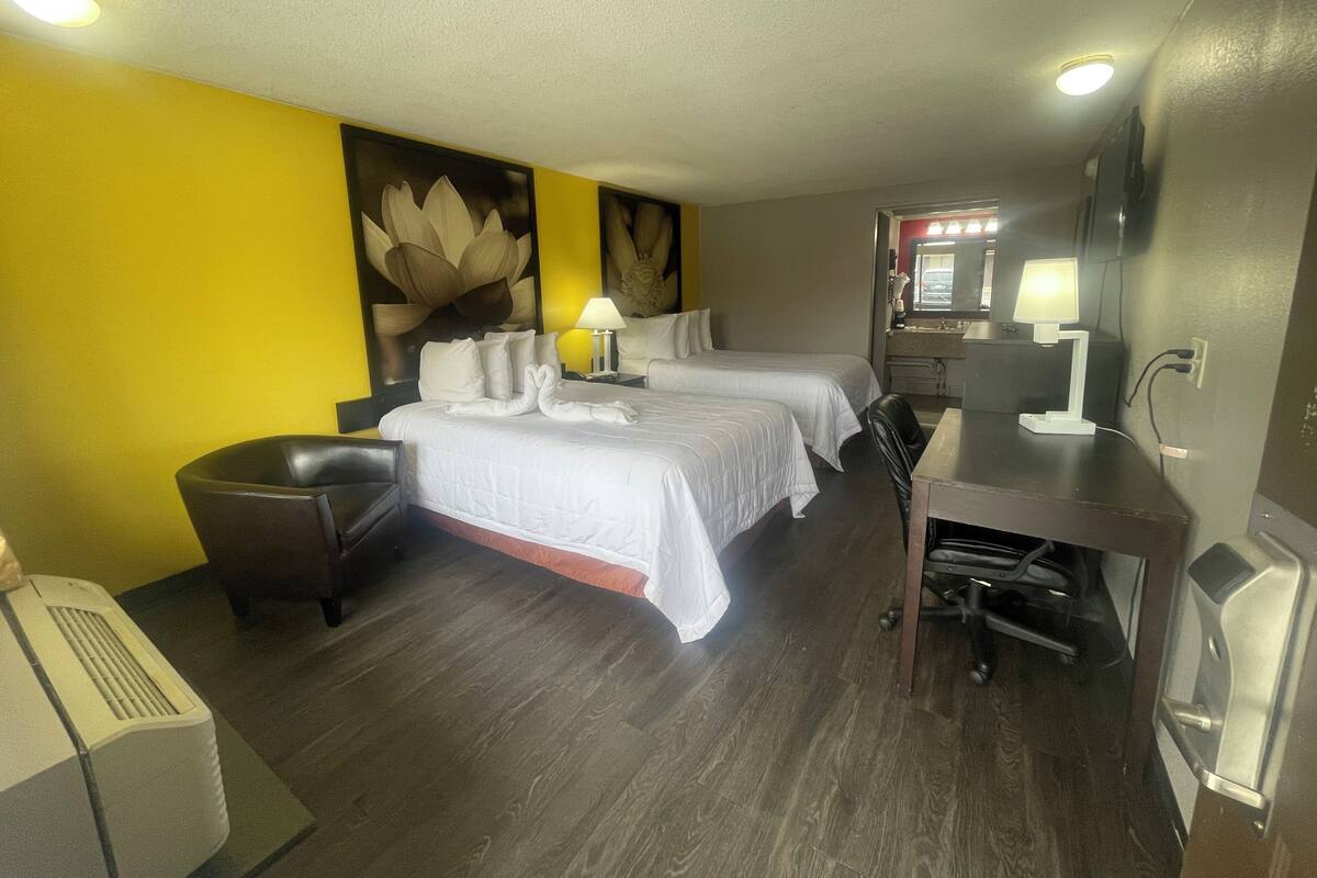 A hotel room with two beds, a desk, a chair, a desk lamp, a mirror, and a wall-mounted air conditioning unit.