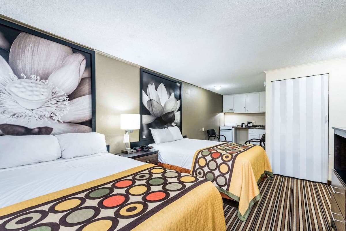 A hotel room with two beds, a large floral artwork on the wall, a kitchenette, and a sliding door.