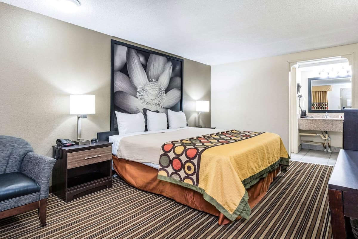 A hotel room with a bed, bedside table, lamp, chair, and a bathroom visible through an open door.
