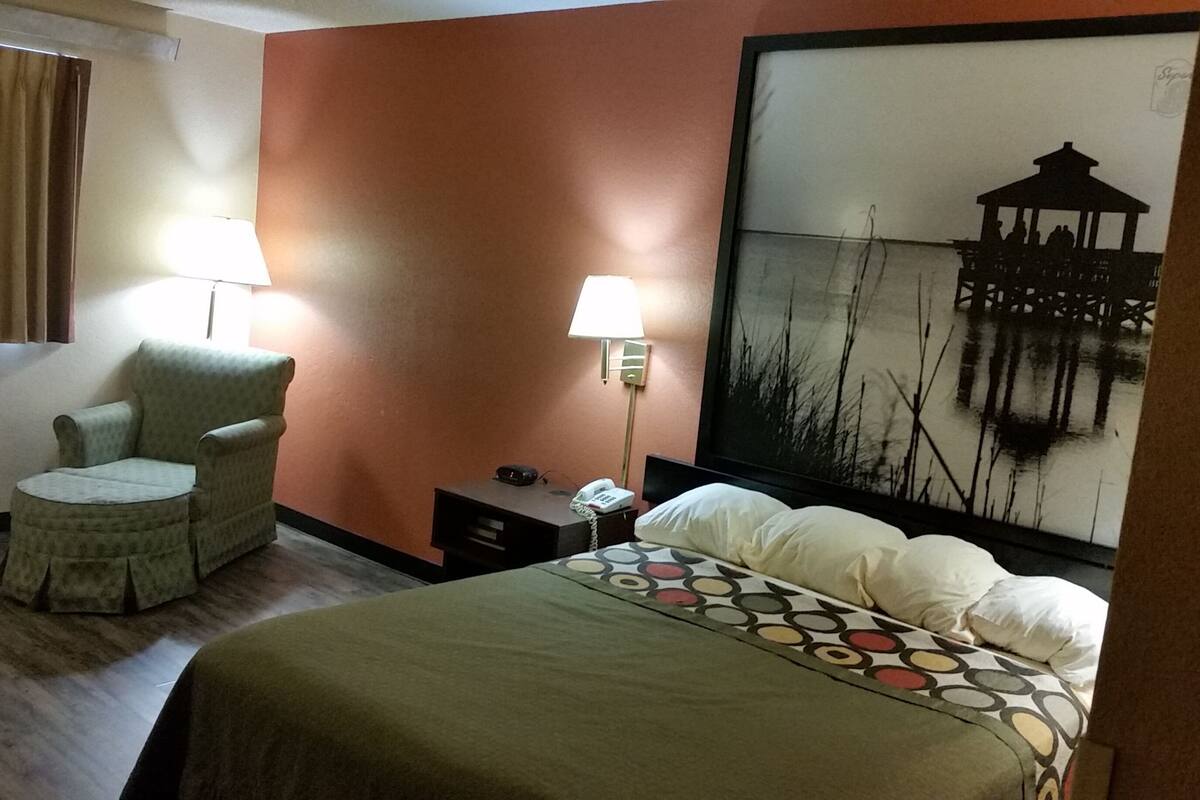 A hotel room with a bed, a chair, a nightstand, a lamp, and a painting on the wall.