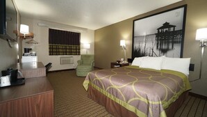 Quarto de hotel com uma cama grande, uma escrivaninha, uma cadeira, um abajur e um quadro na parede.