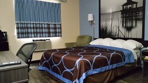 A hotel room with a bed, a desk, a chair, a lamp, and a painting on the wall.