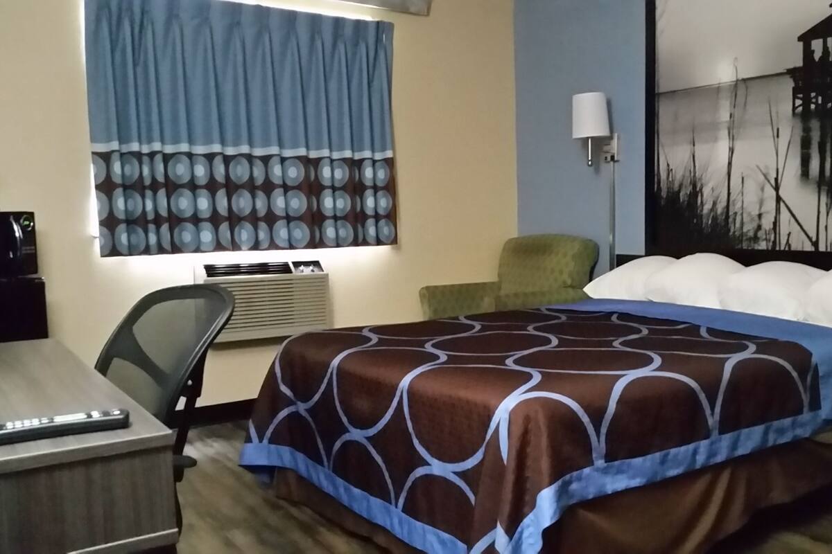 A hotel room with a bed, a desk, a chair, a lamp, and a painting on the wall.