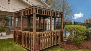 A wooden gazebo with a metal railing, surrounded by a brick building and a pool.