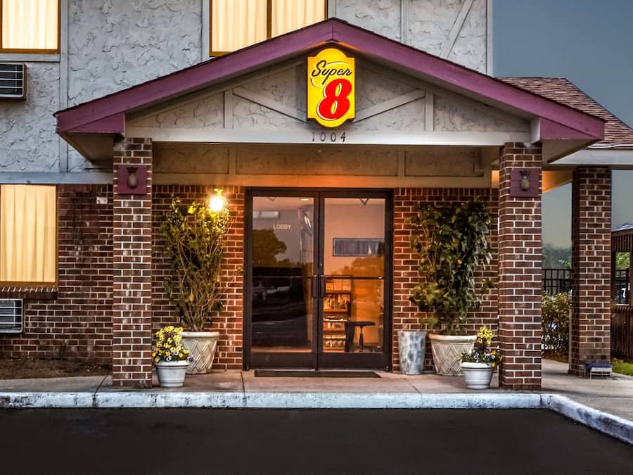Entrada de um hotel com um letreiro chamativo 'Super 8', fachada de tijolos e plantas em vasos.
