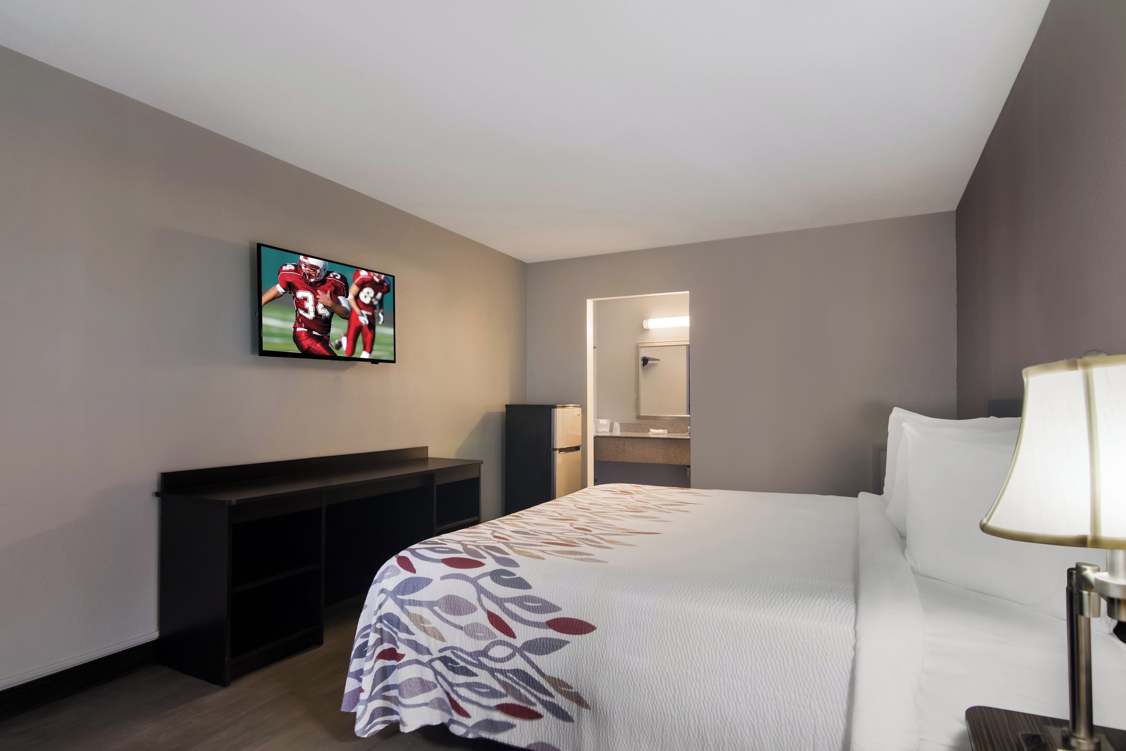 A hotel room with a bed, a TV mounted on the wall, a desk, and a mirror.