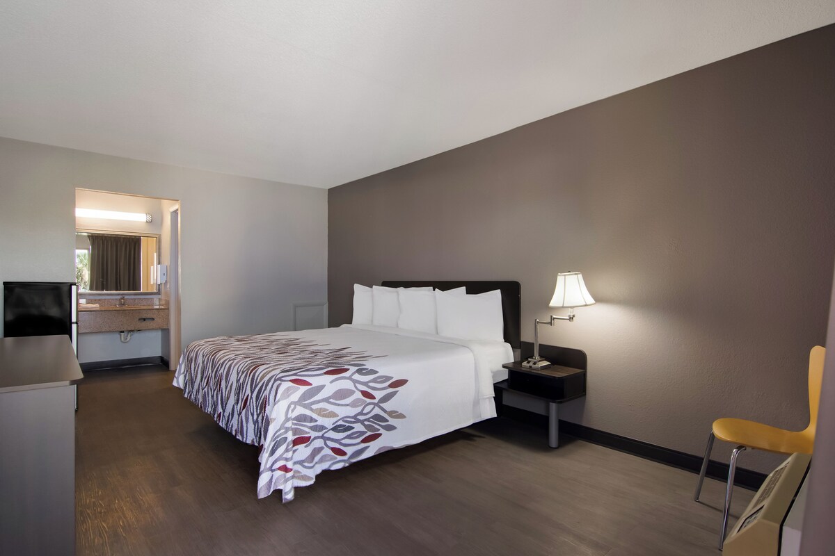 A hotel room with a bed, a desk, a chair, a nightstand with a lamp, and a mirror.