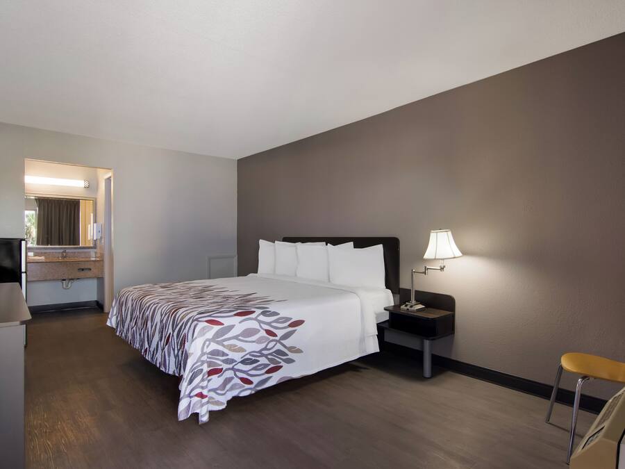 A hotel room with a bed, a desk, a chair, a nightstand with a lamp, and a mirror.