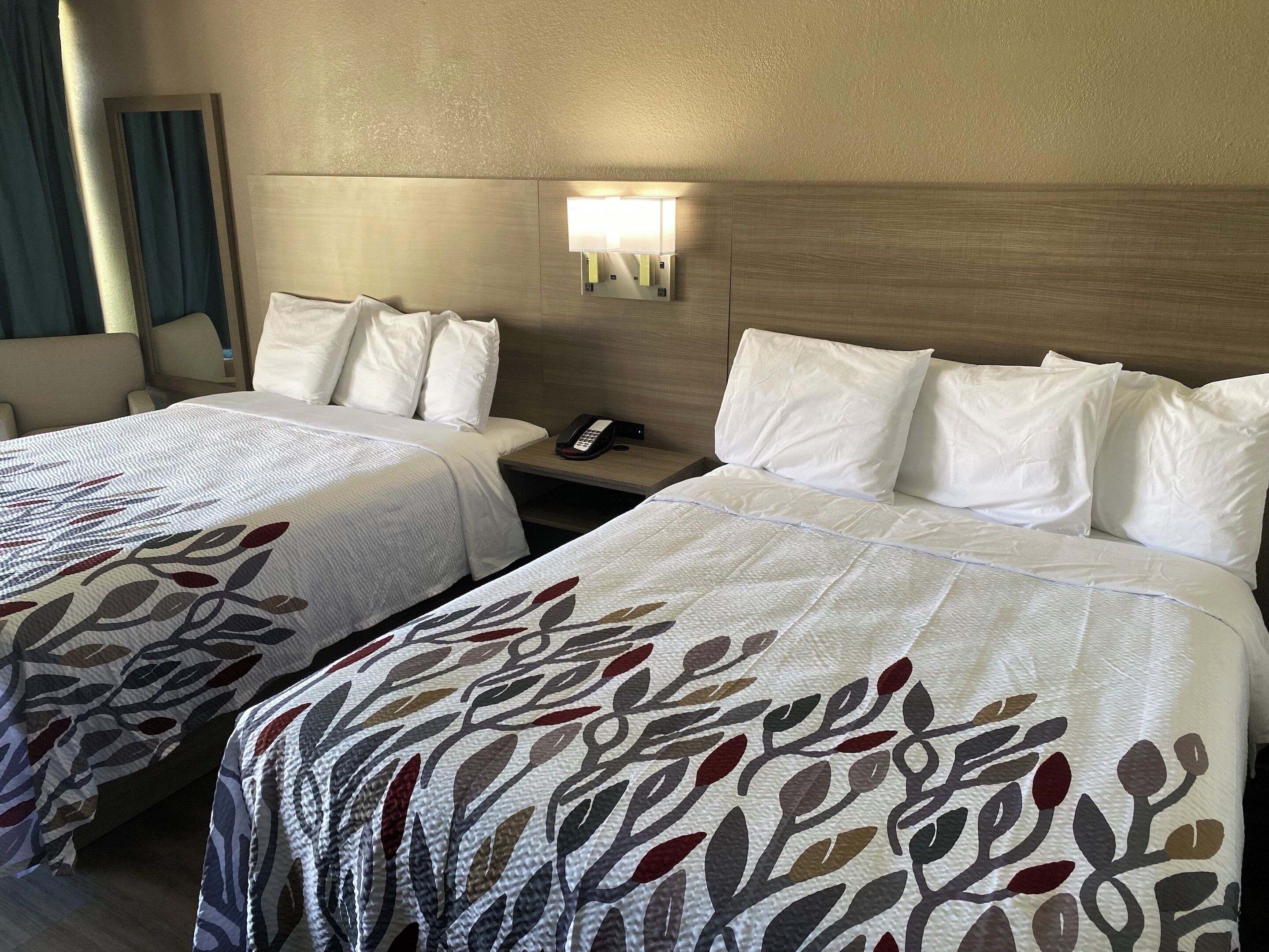 A hotel room with two beds, each with white linens and a patterned blanket. There is a bedside table with a phone and a lamp.