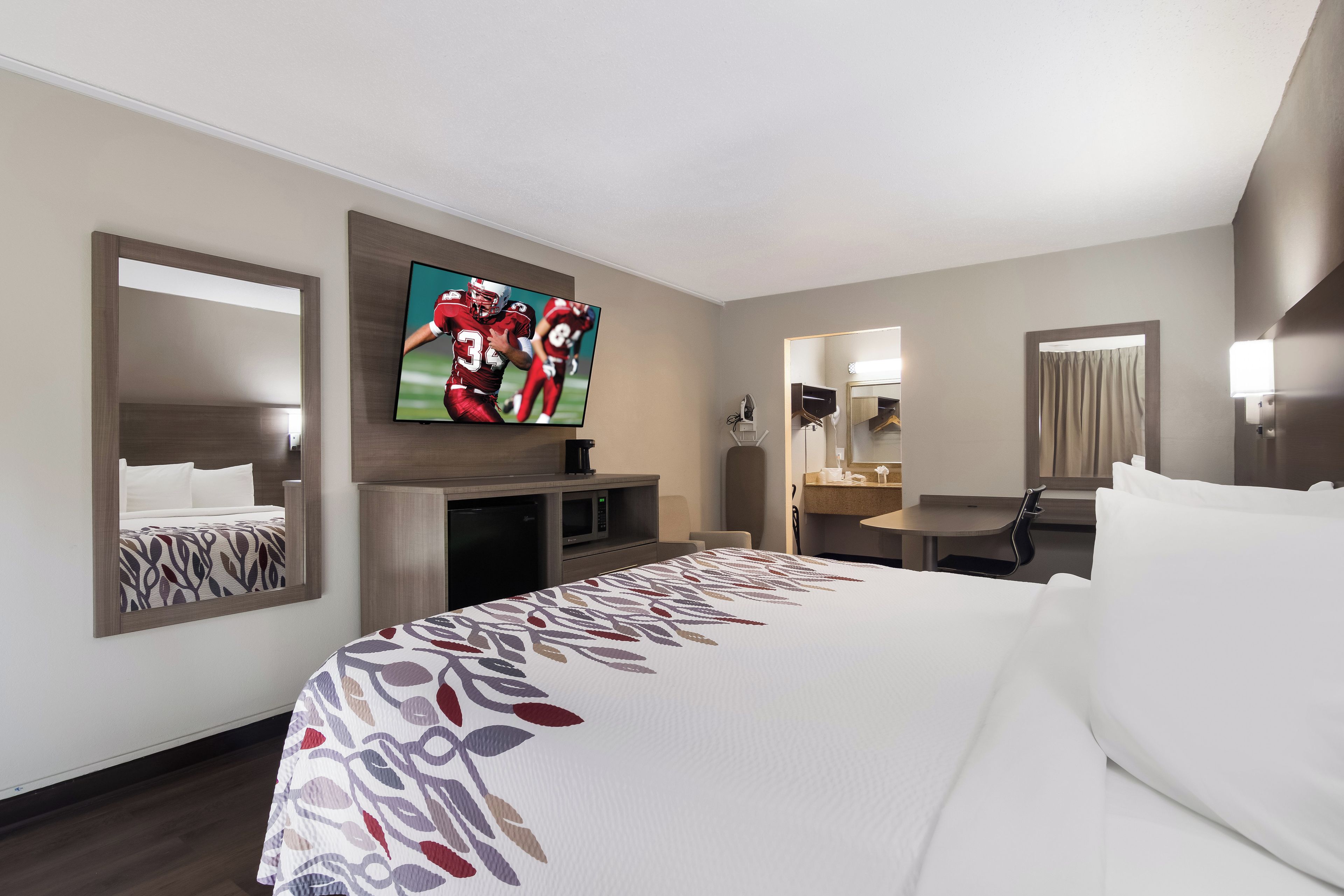 A hotel room with a large bed, a flat-screen TV mounted on the wall, a desk with a chair, a mirror, and a small kitchenette area.