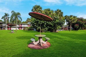 A well-maintained lawn with a central table and umbrella, surrounded by lush greenery and a building with multiple windows in the background. - Red Roof Inn Vero Beach - I-95 (Vero Beach)