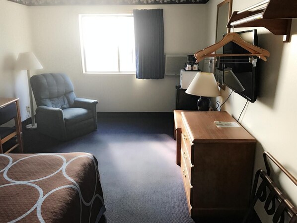 Quarto de hotel com cama, cadeira, escrivaninha, luminária, televisão e janela com cortinas.