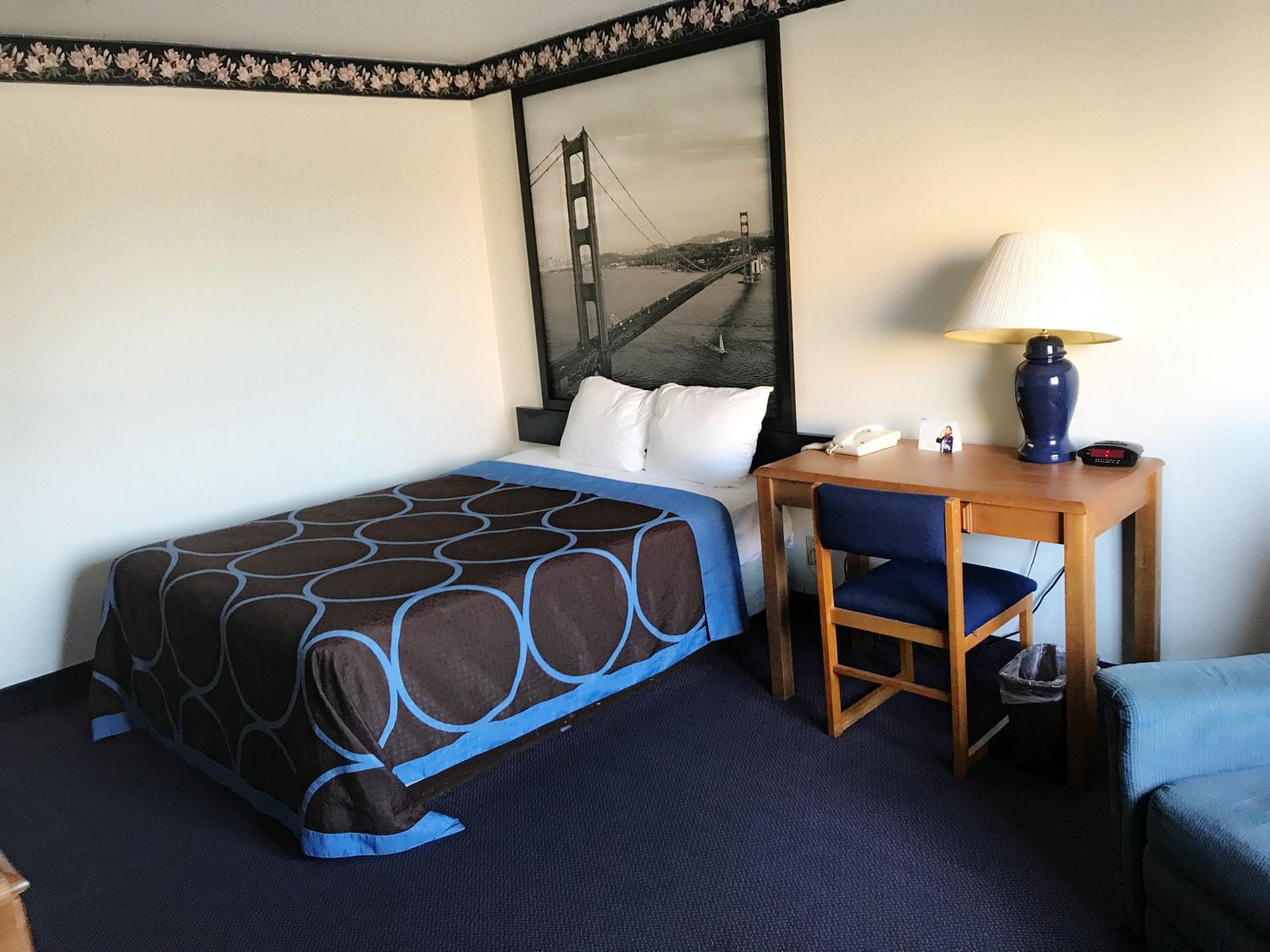 A hotel room with a bed, a desk, a chair, a lamp, and a framed picture of the Golden Gate Bridge.