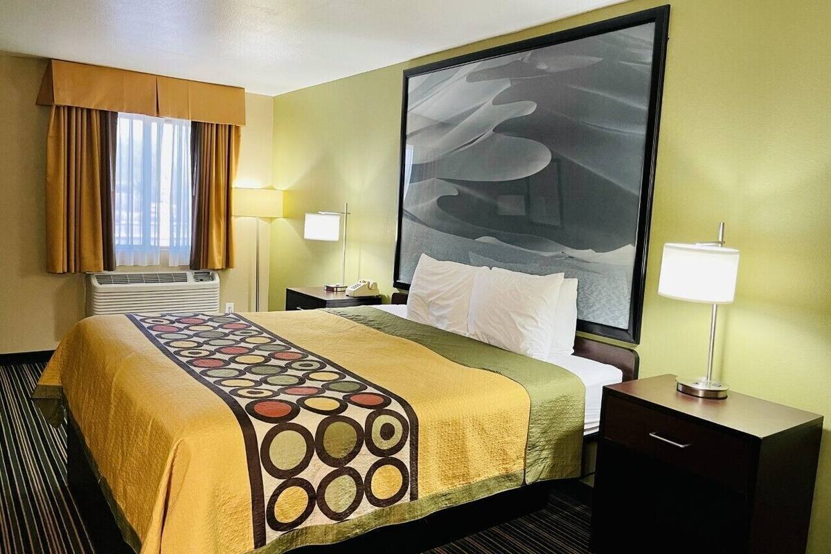 Hotelkamer met een bed, twee nachtkastjes, een groot raam met gordijnen, een wandairconditioner en een schilderij.