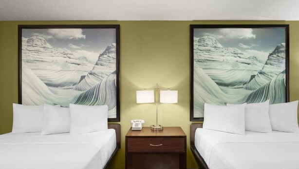 Une chambre d’hôtel avec deux lits, une table de chevet avec une lampe et deux tableaux représentant des paysages, encadrés et accrochés aux murs.