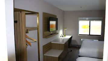 A hotel room with a wooden wardrobe, a flat-screen TV mounted on the wall, a bedside table with a lamp, and a window with a view of greenery.