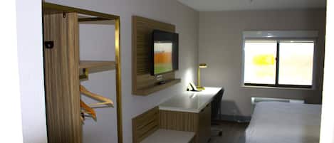 A hotel room with a wooden wardrobe, a flat-screen TV mounted on the wall, a bedside table with a lamp, and a window with a view of greenery.