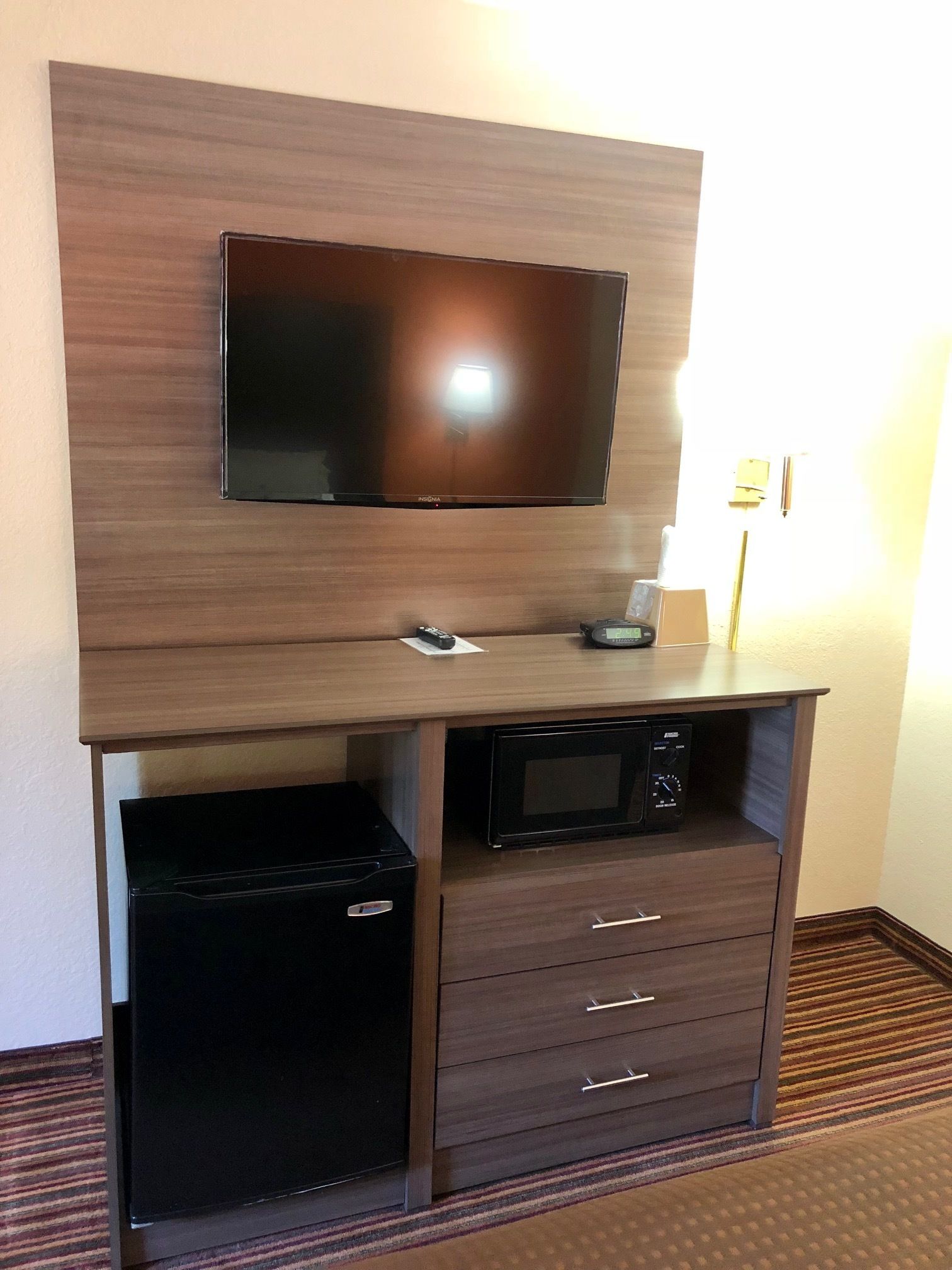 Een compacte hotelkamer met een flatscreen televisie bevestigd aan een houten kast, een kleine koelkast en een magnetron.