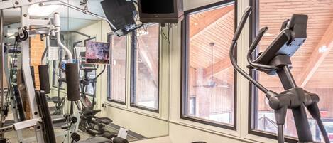 Un gimnasio con equipo de ejercicio, ventanas amplias y una televisión montada en el techo.