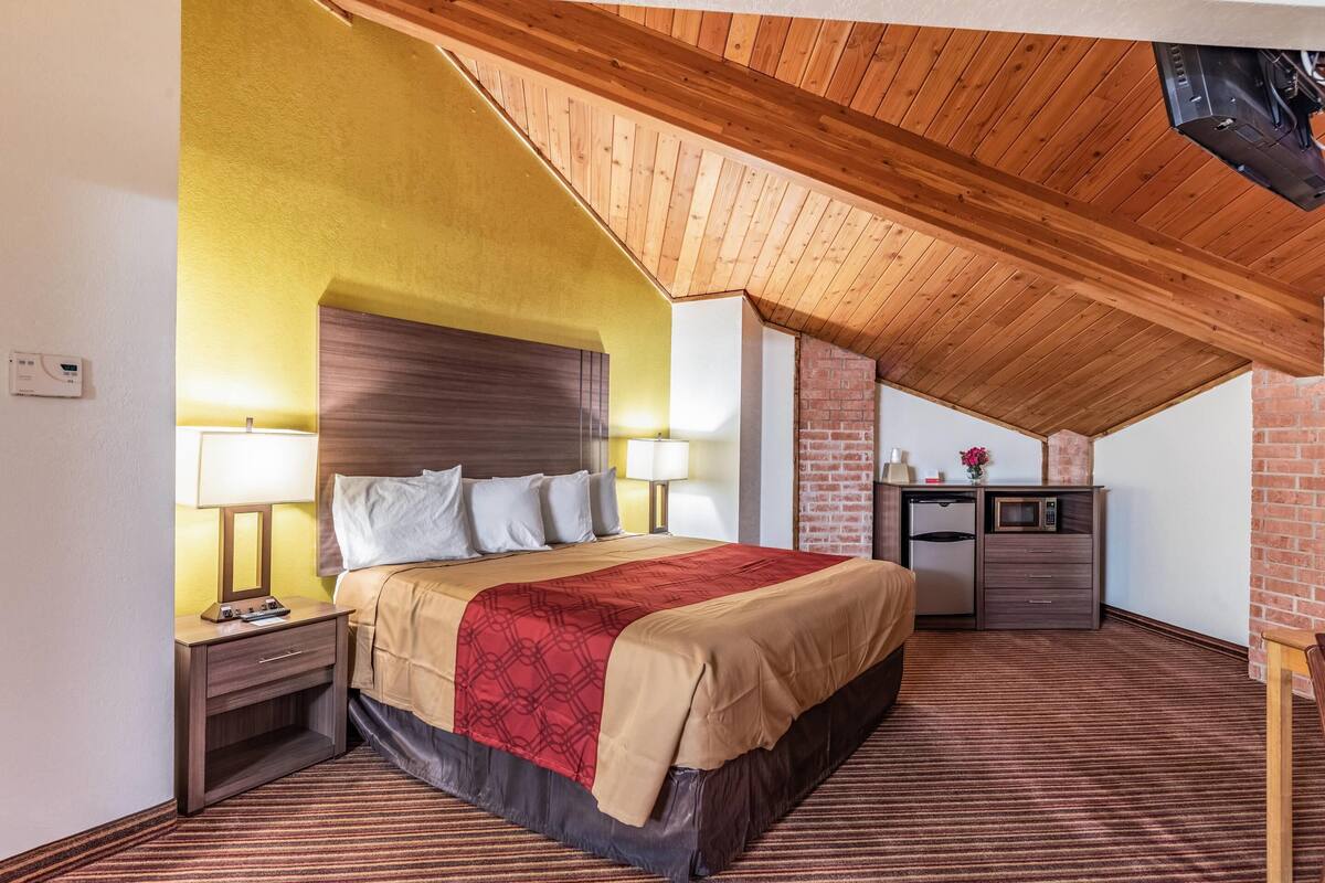 A hotel room with a wooden ceiling, a bed with a red and beige bedspread, a nightstand with a lamp, a desk with a microwave, and a brick wall.
