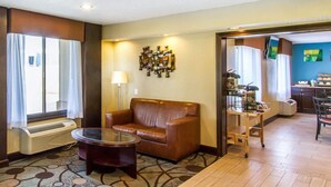 A hotel room with a brown leather sofa, a glass coffee table, a patterned rug, a wall-mounted air conditioner, and a view of the dining area through an open doorway. - Quality Inn Murray University Area (Murray)