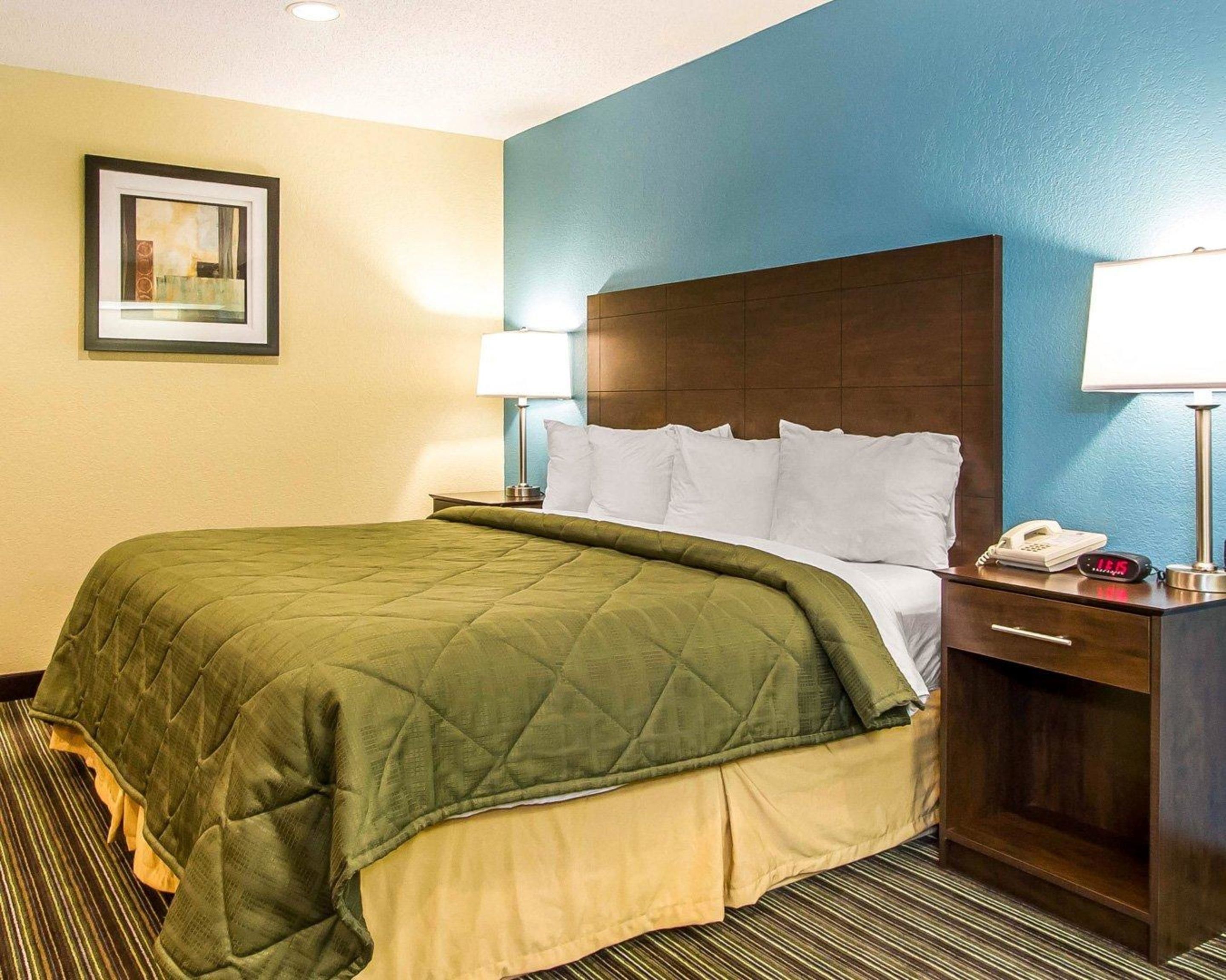 A hotel room with a bed, a nightstand with a lamp, a phone, and a digital clock.