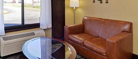 A hotel room with a leather sofa, a glass-top coffee table, a standing lamp, and a view of a parking lot through a large window.