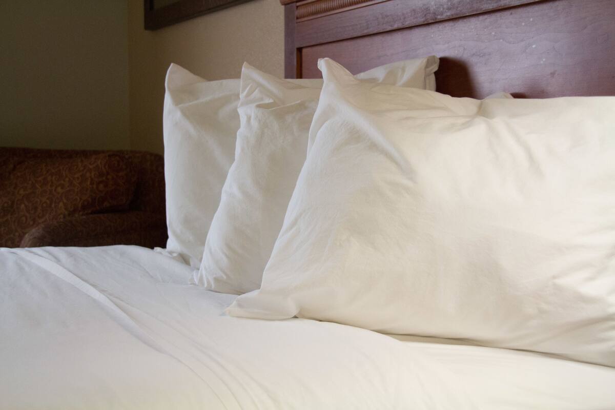 Una cama bien hecha con ropa de cama y almohadas blancas, un cabecero de madera y un sofá marrón con estampado al fondo.