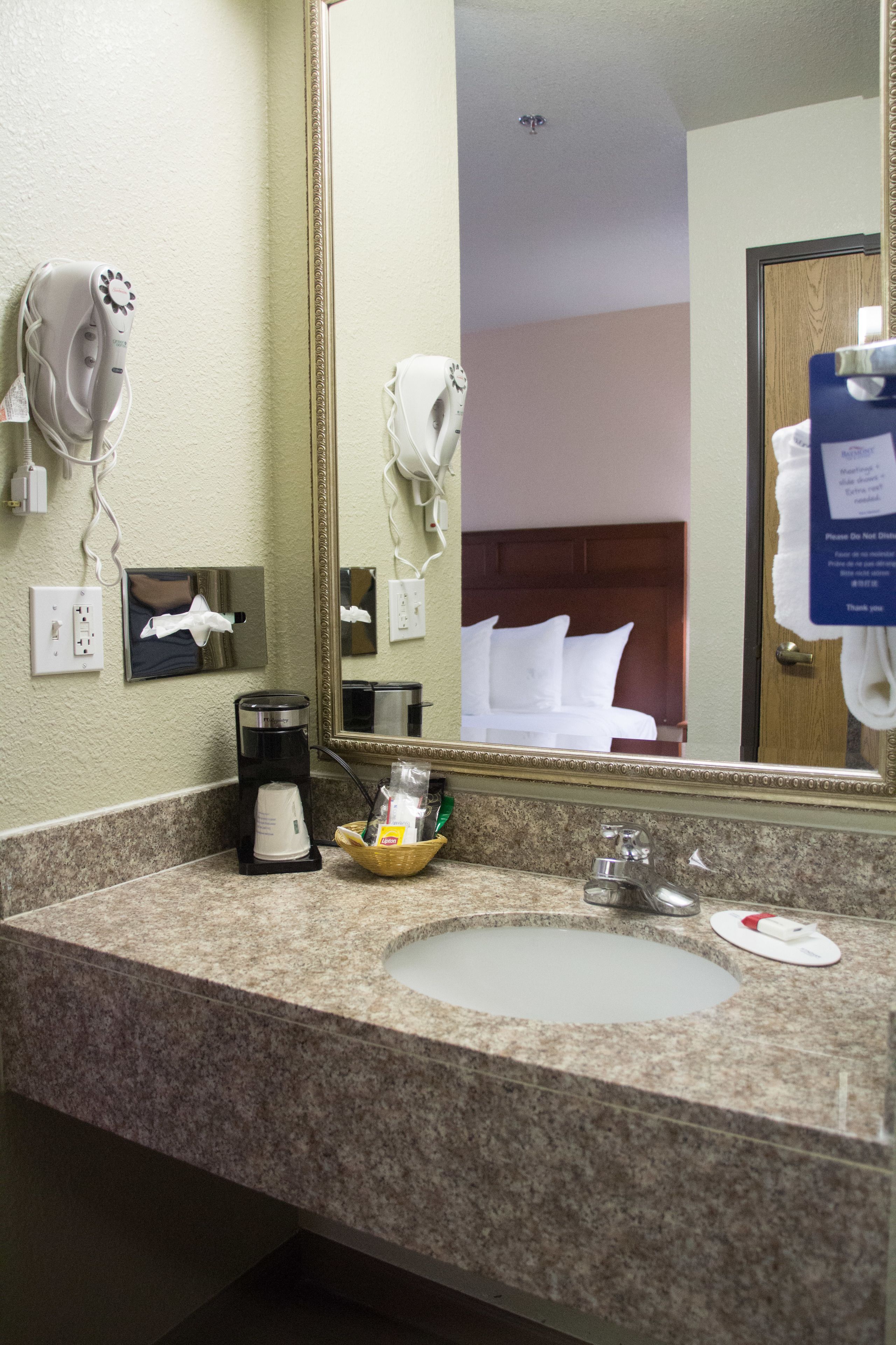 A hotel bathroom with a granite countertop, a coffee maker, a hand dryer, and a mirror reflecting a bed and a door.