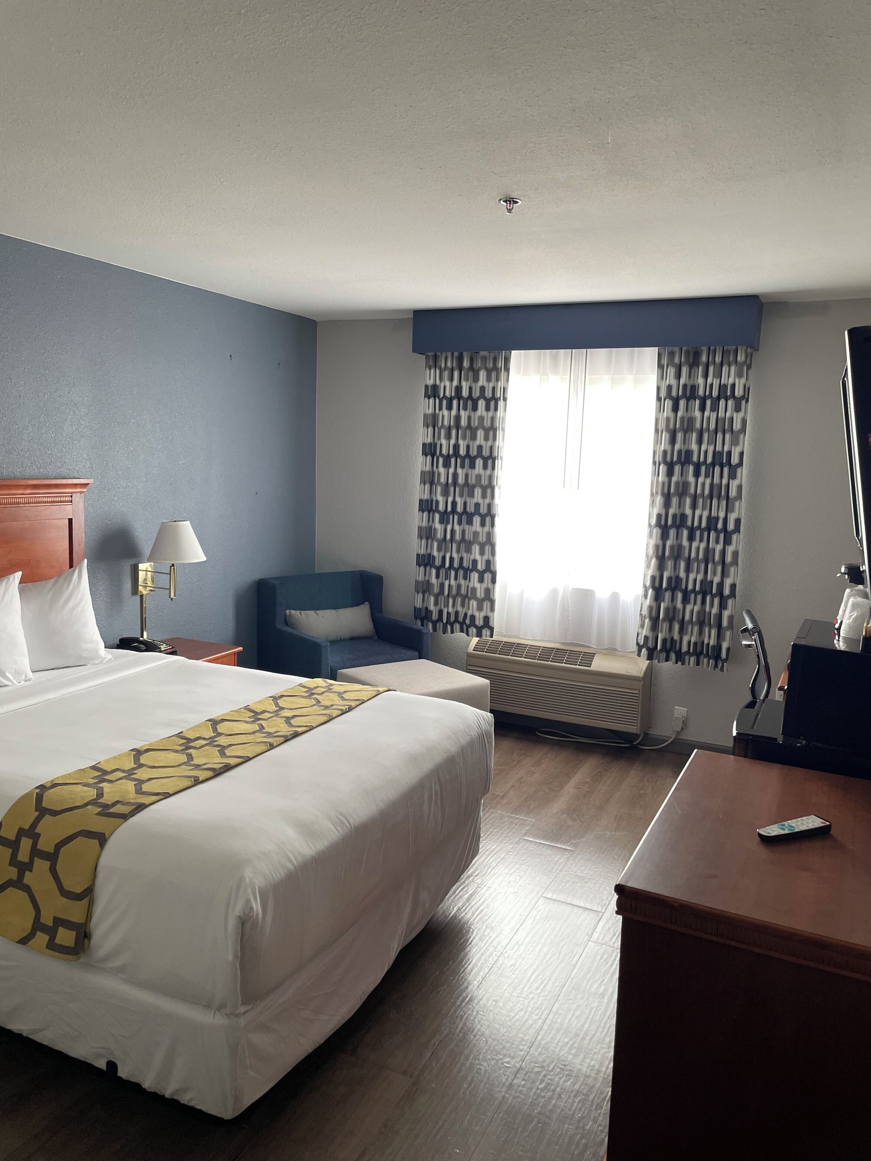 A hotel room with a large bed, a desk, a chair, a desk lamp, an air conditioning unit, and a window with patterned curtains.