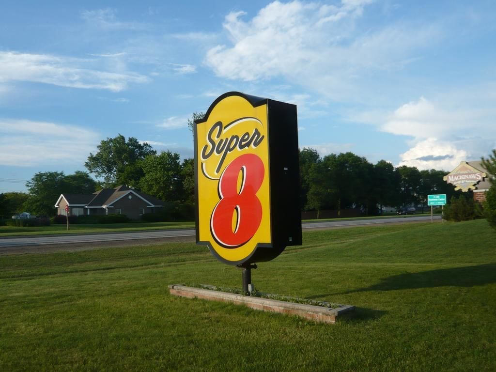 A large, bold sign with the text "Super 8" in a yellow and red color scheme, set against a backdrop of a grassy area with a road and houses in the distance.