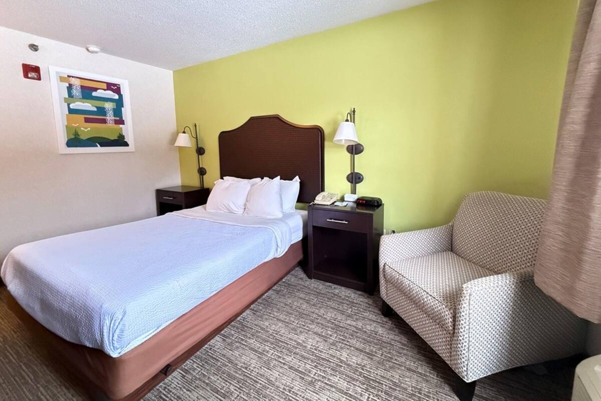 A hotel room with a bed, a chair, a nightstand, and a wall-mounted lamp.