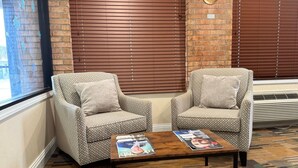 Two patterned armchairs with cushions, a wooden coffee table with magazines, and a brick wall with a window and blinds. - SureStay by Best Western Grove City (Grove City)