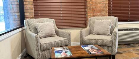 Two patterned armchairs with cushions, a wooden coffee table with magazines, and a brick wall with a window and blinds.