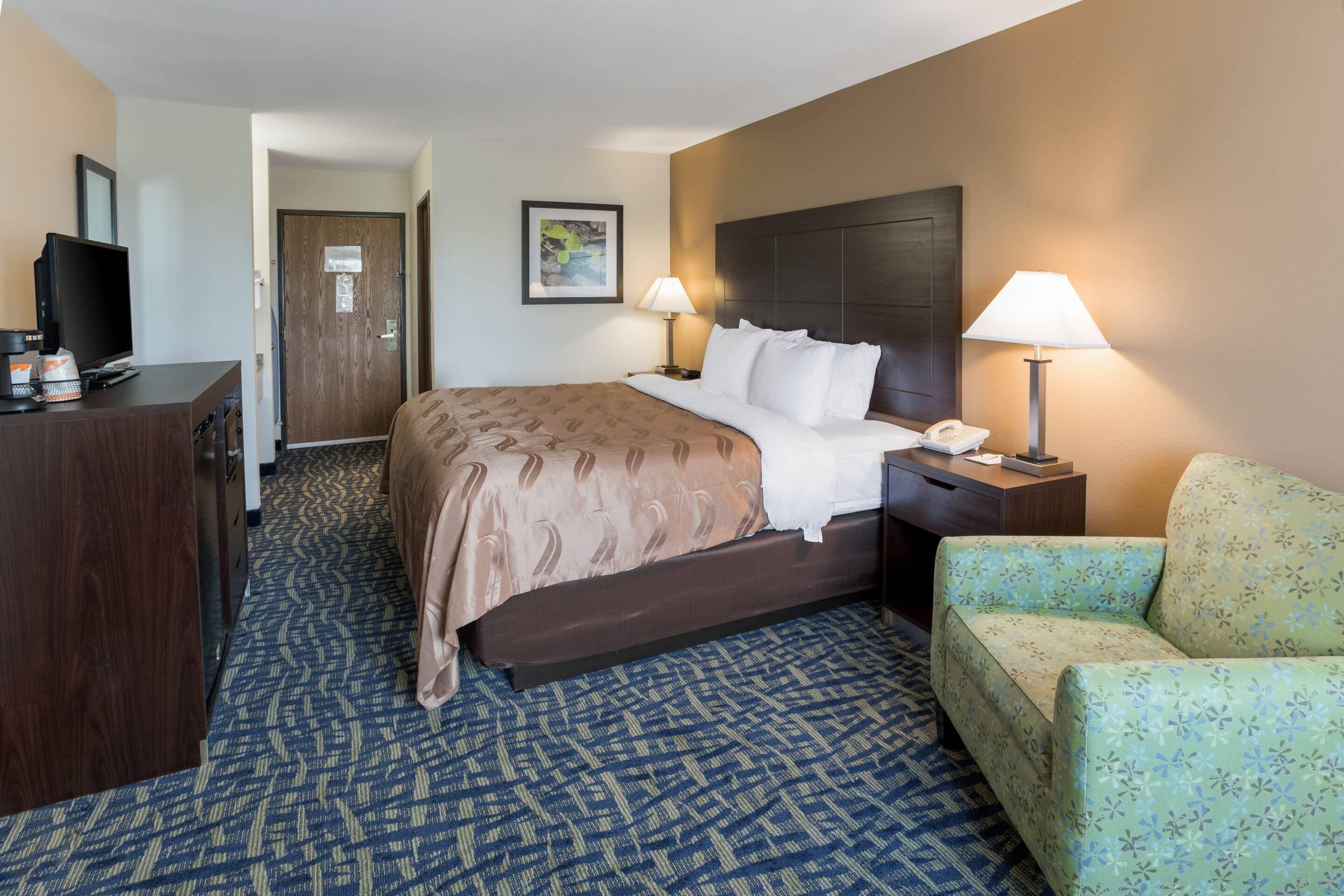 A hotel room with a bed, bedside lamps, a desk with a television, a chair, and a door.