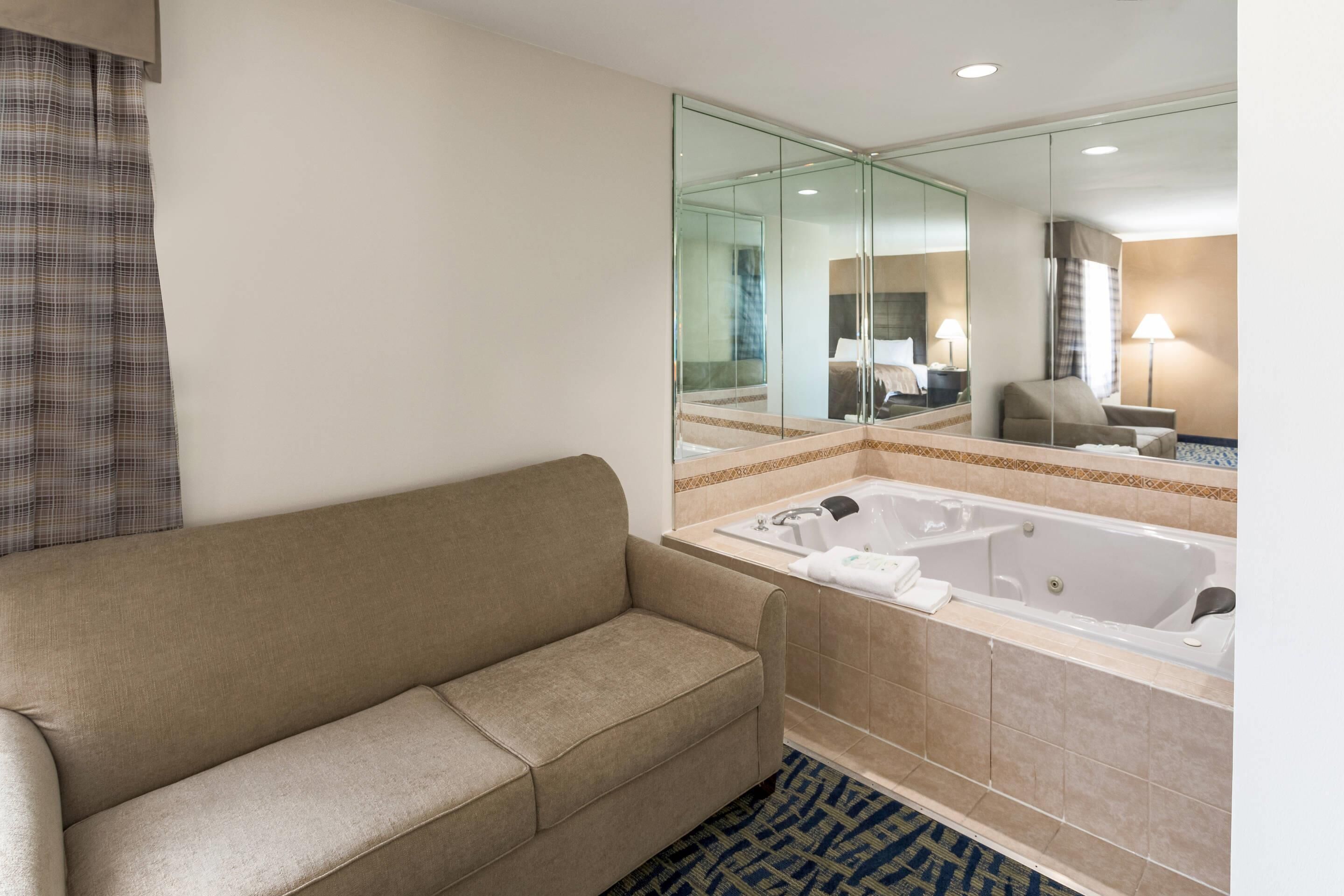A hotel bathroom with a beige sofa, a large mirror, a bathtub, and a patterned rug.