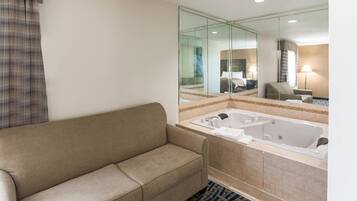 A hotel bathroom with a beige sofa, a large mirror, a bathtub, and a patterned rug.