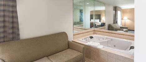 Un baño de hotel con un sofá color beige, un espejo grande, una bañera y un tapete estampado.