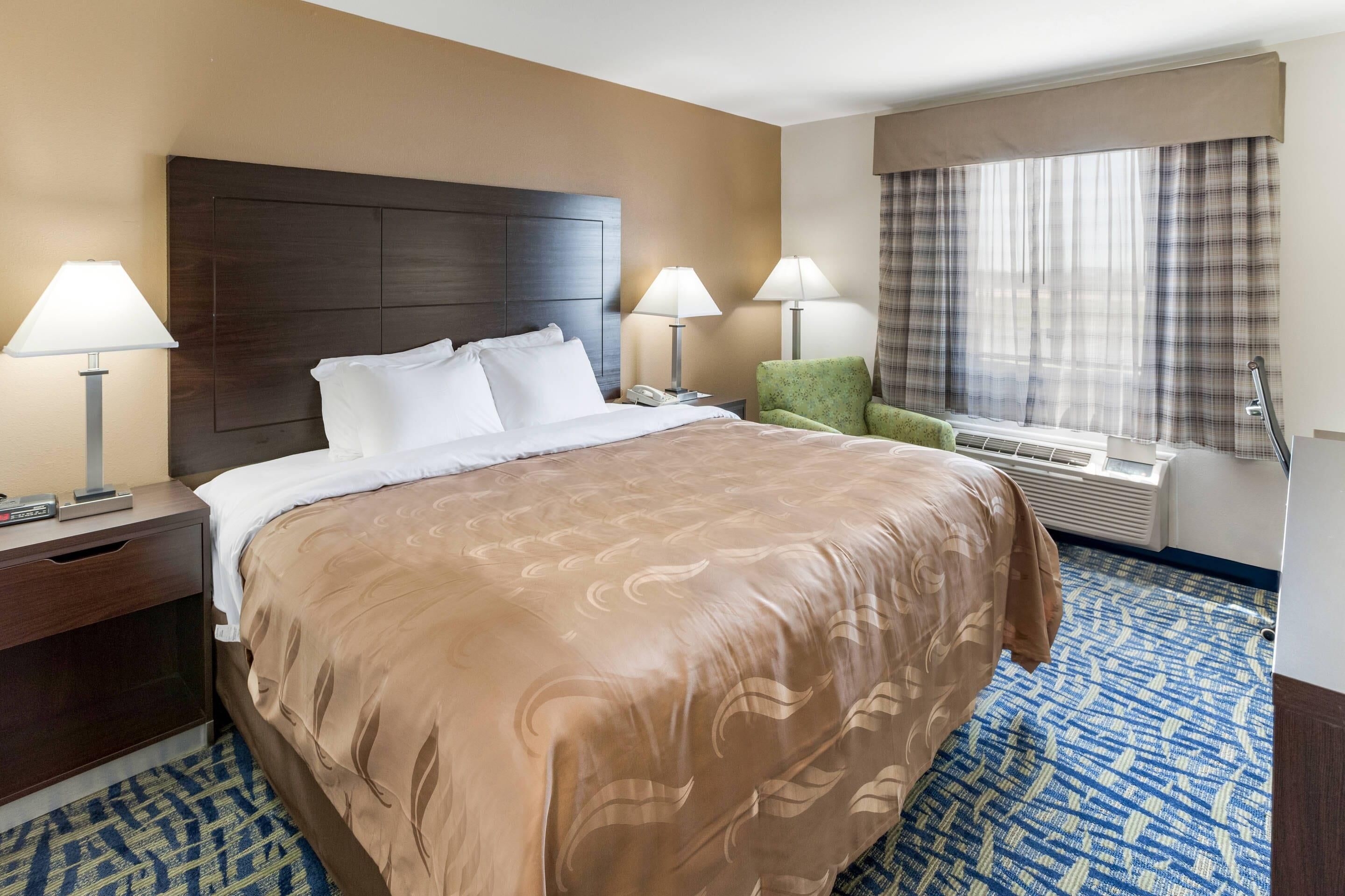 A hotel room with a large bed, two bedside lamps, a green chair, a window with curtains, and an air conditioning unit.